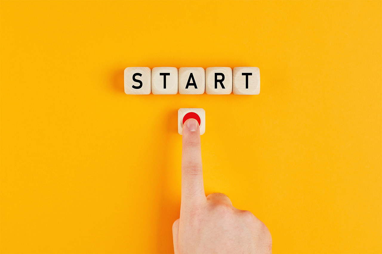 黄色い背景にウッドブロックで書かれた「Start」の文字。その下側に赤いボタンがあり、人差し指で推されている。